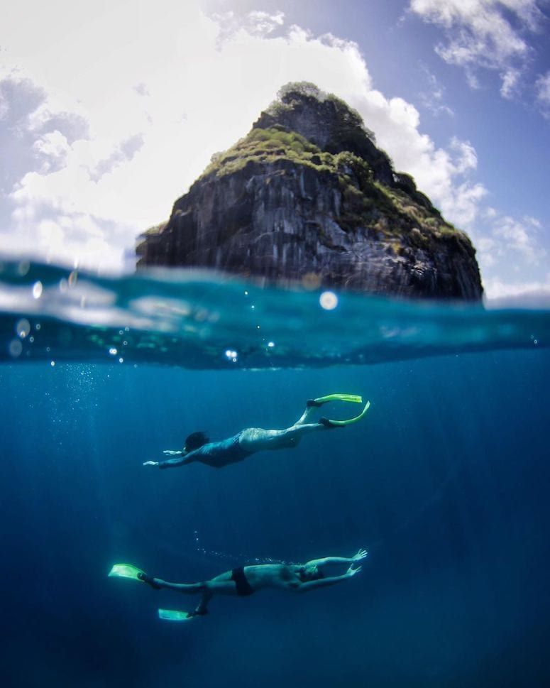 Mergulhadores em apneia sob a água com o Morro do Pico ao fundo em Fernando de Noronha